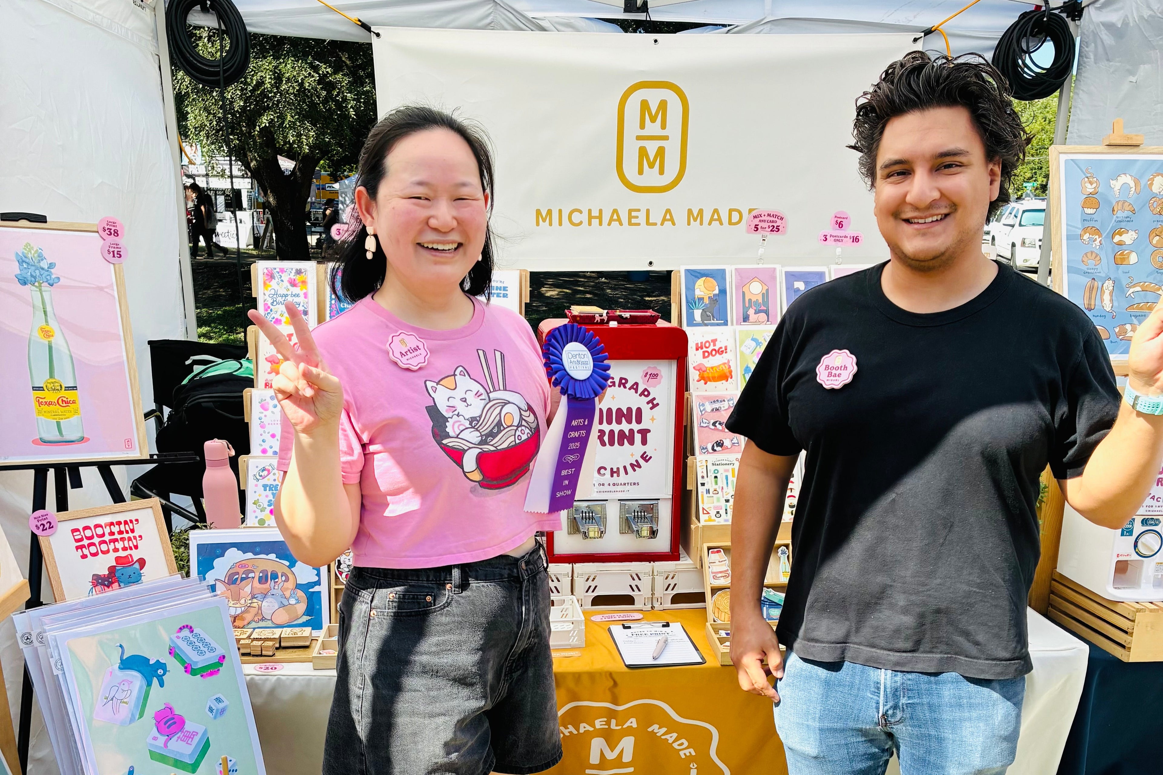 Two people standing in front of a booth with 'Michaela Mag' branding at an outdoor event.